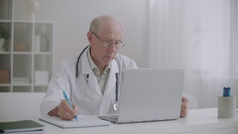 Der-ältere-Arzt-Hört-Dem-Patienten-Bei-Der-Online-Konsultation-Zu,-Schaut-Auf-Den-Bildschirm-Des-Laptops-Und-Schreibt-Notizen-Auf-Papier